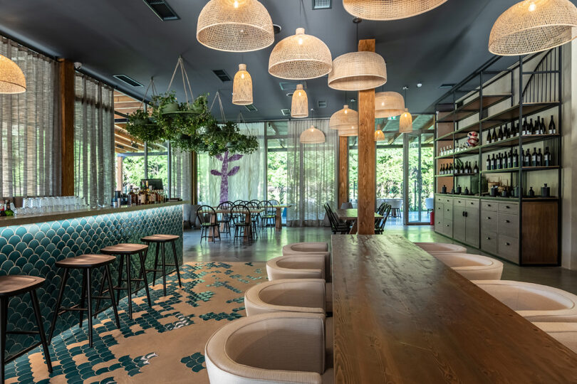 The modern restaurant interior at Lopota Lake Resort features a long wooden table, cushioned chairs, and a stylish bar area. Pendant lights elegantly hang from the ceiling, while shelves adorned with bottles and lush greenery create an inviting backdrop.