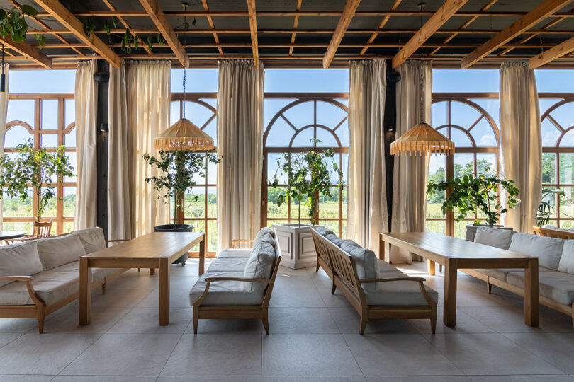 Spacious room with large arched windows, wooden ceiling beams, and minimalist furniture reminiscent of Lopota Lake Resort. Indoor plants adorn the space, and sunlight streams through sheer curtains.