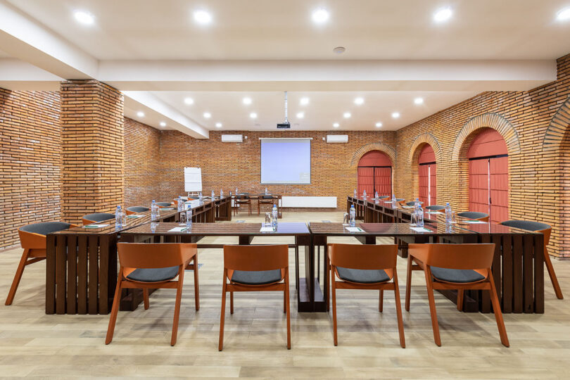 Conference room at Lopota Lake Resort featuring a U-shaped table setup, brown chairs, brick walls, wooden floor, ceiling lights, and a projection screen at the front.