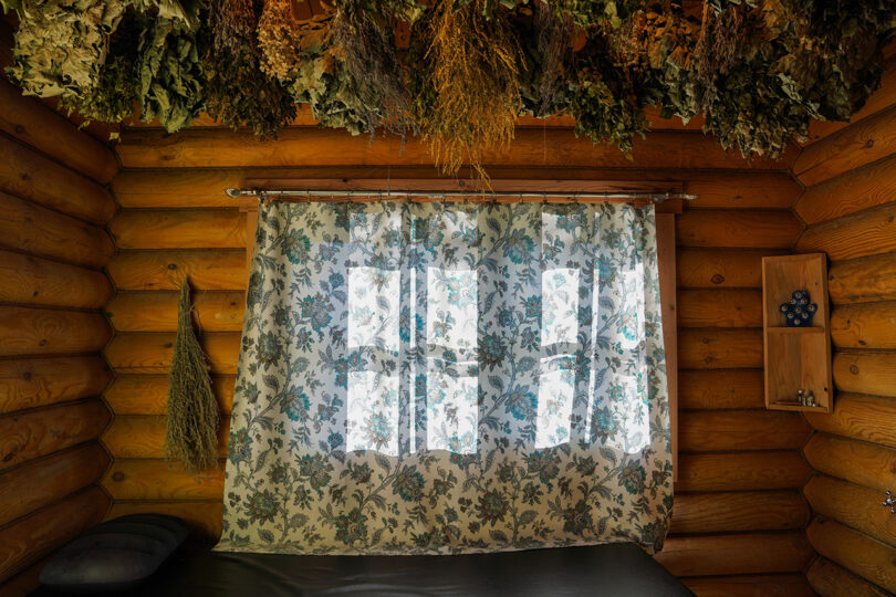 A rustic log cabin interior at Lopota Lake Resort features a floral curtain covering a window, herbs hanging from the ceiling, and a small shelf with four blue stones on the right wall.