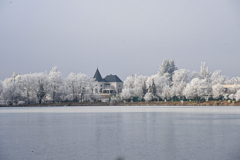 A snow-covered landscape features a house with a pointed roof, nestled among frost-laden trees at Lopota Lake Resort, all beautifully reflected in the calm, frozen lake.