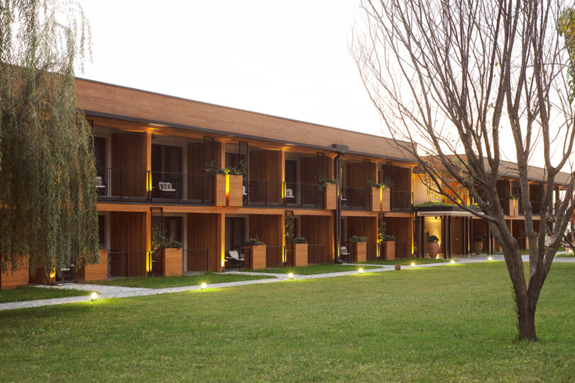 The two-story building at Lopota Lake Resort features a charming wooden exterior and inviting balconies, perfectly framed by the warm hues of sunset. A gently lit pathway meanders through the grass lawn, creating a serene ambiance that welcomes you to this tranquil escape.