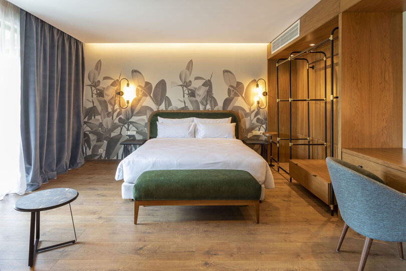 Experience the tranquility of a contemporary hotel room reminiscent of Lopota Lake Resort, featuring a large bed with a green headboard, wooden flooring, botanical wallpaper, modern light fixtures, a desk and chair, and an open closet.