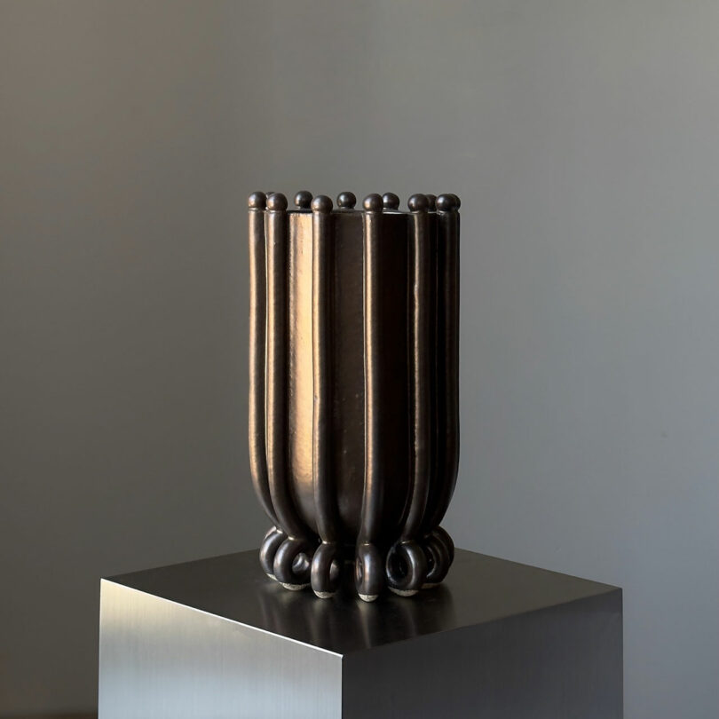 A tall, dark vase with vertical ridges and a rounded base sits on a metallic pedestal against a plain background.