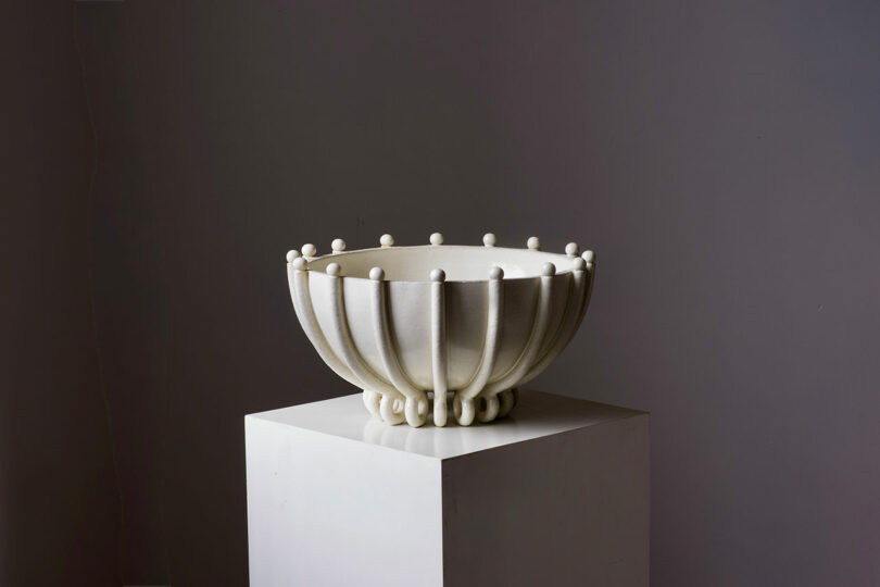 A white, intricately designed ceramic bowl with a textured surface and rounded protrusions sits on a white pedestal against a plain gray background.