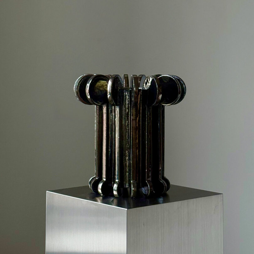 A metallic, cylindrical sculpture with rounded ends is placed on a polished pedestal against a neutral background.