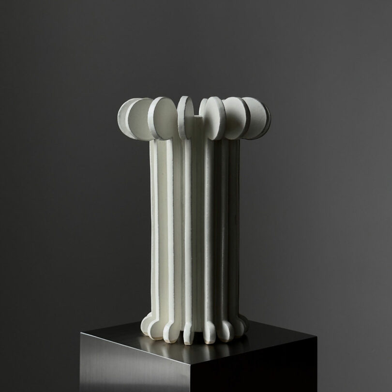 A modern, abstract white sculpture composed of vertical cylindrical elements, topped with circular shapes, displayed on a dark pedestal against a gray background.