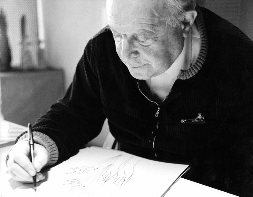 An older man with a pen writes on a paper pad at a desk, deeply focused on his work, wearing a zip-up sweater. His style echoes the timeless elegance of Gio Ponti, blending functionality with artistic flair.