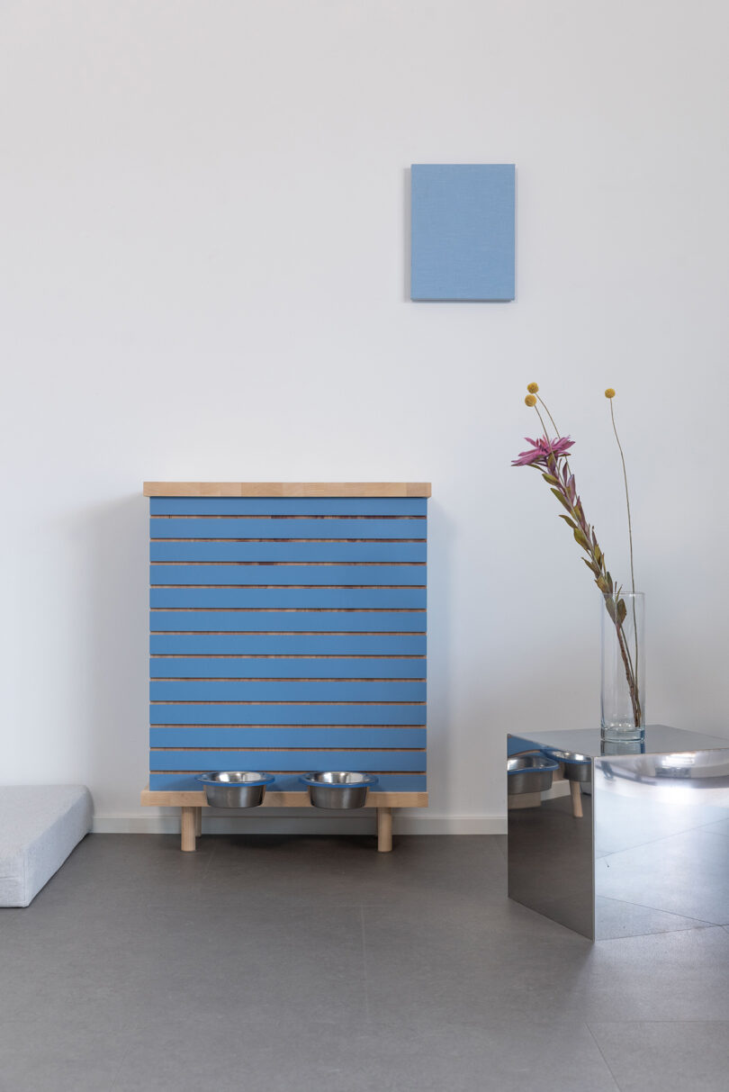 Minimalist room with a blue-striped cabinet holding pet bowls, a small blue wall panel above, a tall flower in a glass vase on a mirrored table, and a light gray floor cushion.