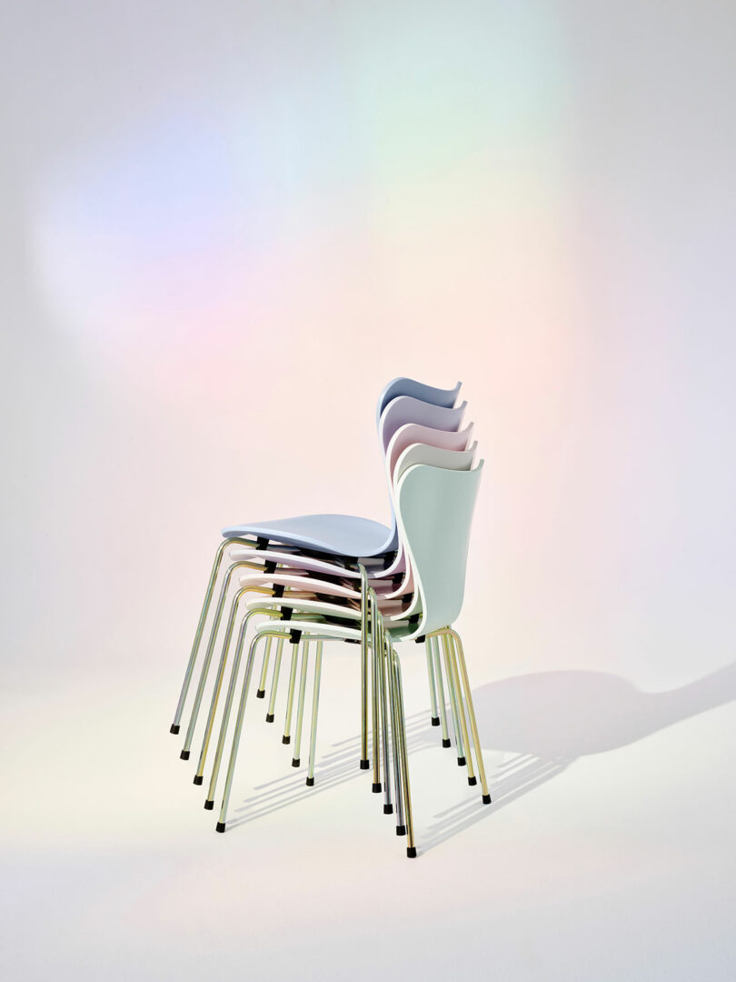 A stack of six modern chairs with sleek metal legs and pastel-colored seats, arranged against a softly lit background.