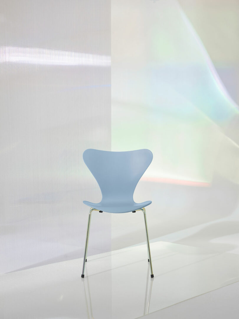 A light blue chair with a curved backrest and silver legs is placed in a minimalist setting with soft, iridescent lighting.