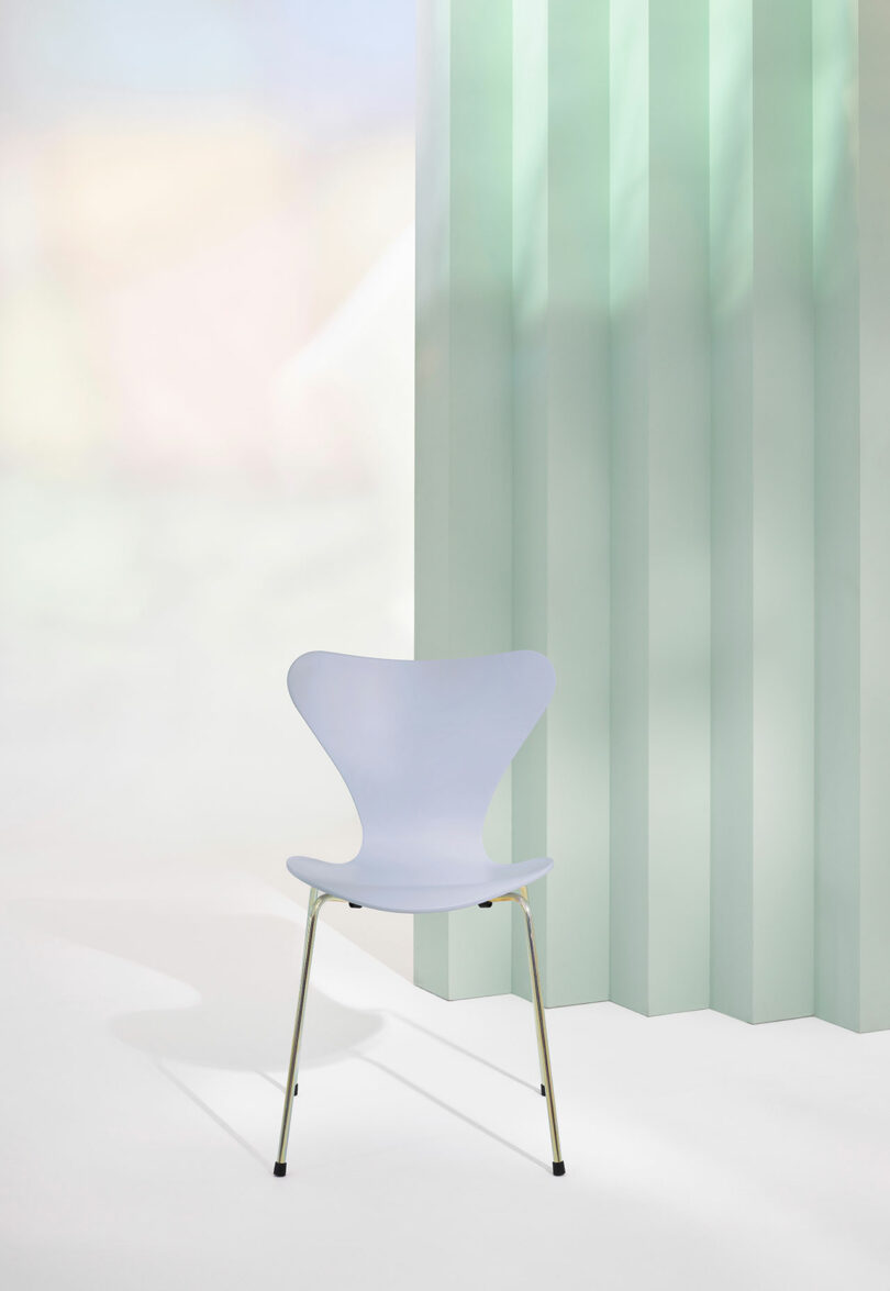 A modern, minimalist chair with a light blue seat and metal legs is placed on a white floor next to a mint green, vertically striped wall.