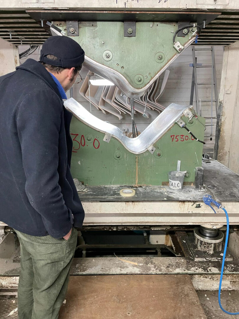 A person in a black jacket and cap observes a large industrial machine pressing metal sheets.