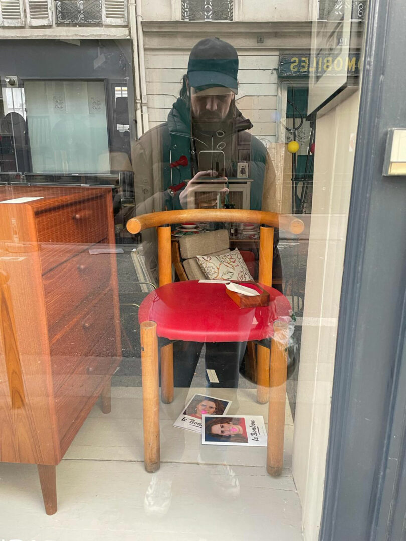 Person taking a selfie in a storefront window reflection, with a red chair and wooden dresser visible inside. Flyers with faces are on the floor beneath the chair.