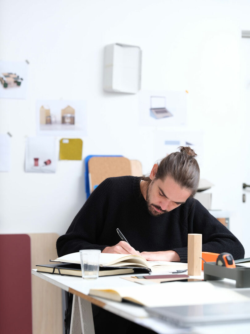 A person with long hair tied back is writing in a notebook at a desk. The desk has another open notebook, a glass, and office items. Papers are on the white wall in the background.