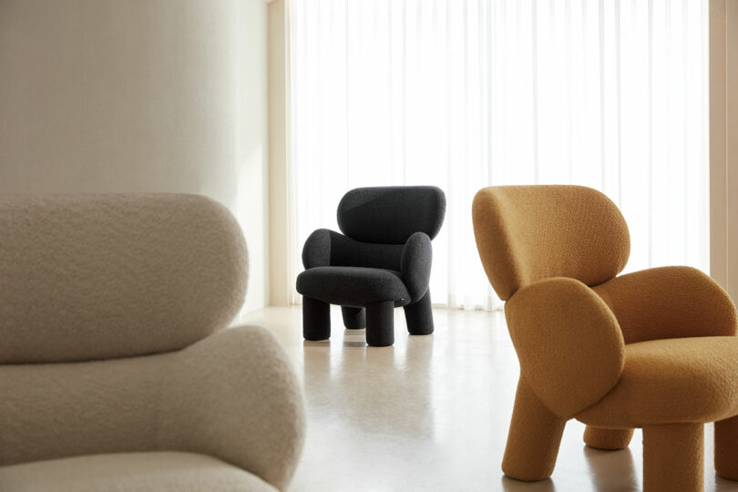 Three modern, textured armchairs in beige, black, and mustard are positioned across a neutral, light-filled room with sheer curtains.