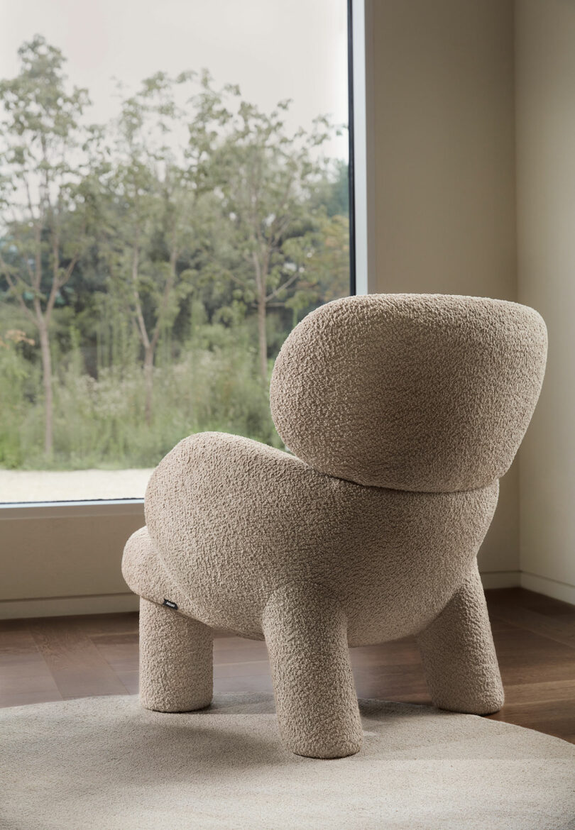 Beige, plush armchair with rounded design placed by a large window overlooking a garden with trees.