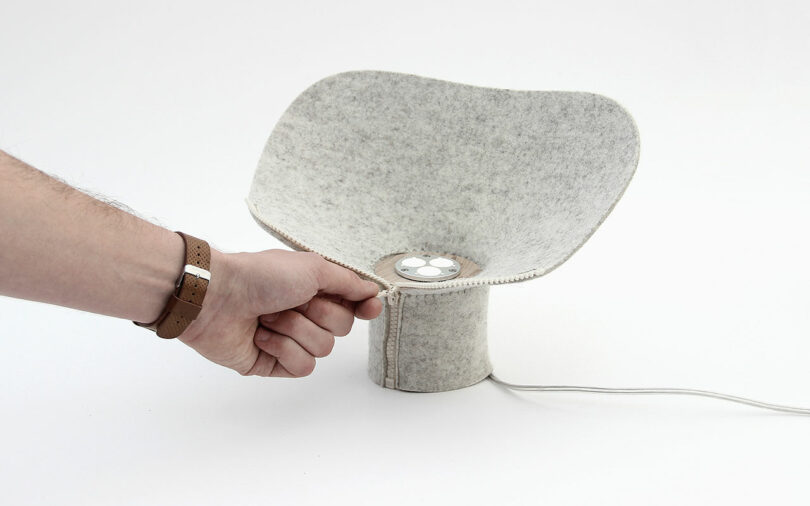 A hand adjusts a gray felt lampshade with a wooden base. The lamp has a white cord extending from its base.