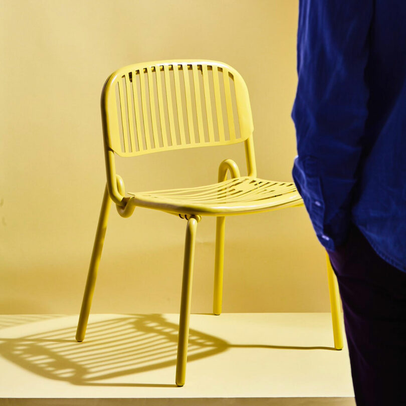 A yellow slatted chair sits on a yellow platform with shadows cast beneath. A person in a blue shirt is partially visible in the foreground.