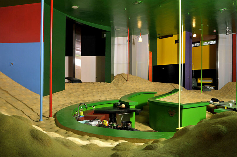 A room with wavy, sand-like flooring and colorful geometric walls. A kitchen area with bright green counters is central, surrounded by yellow mounds and vertical poles.