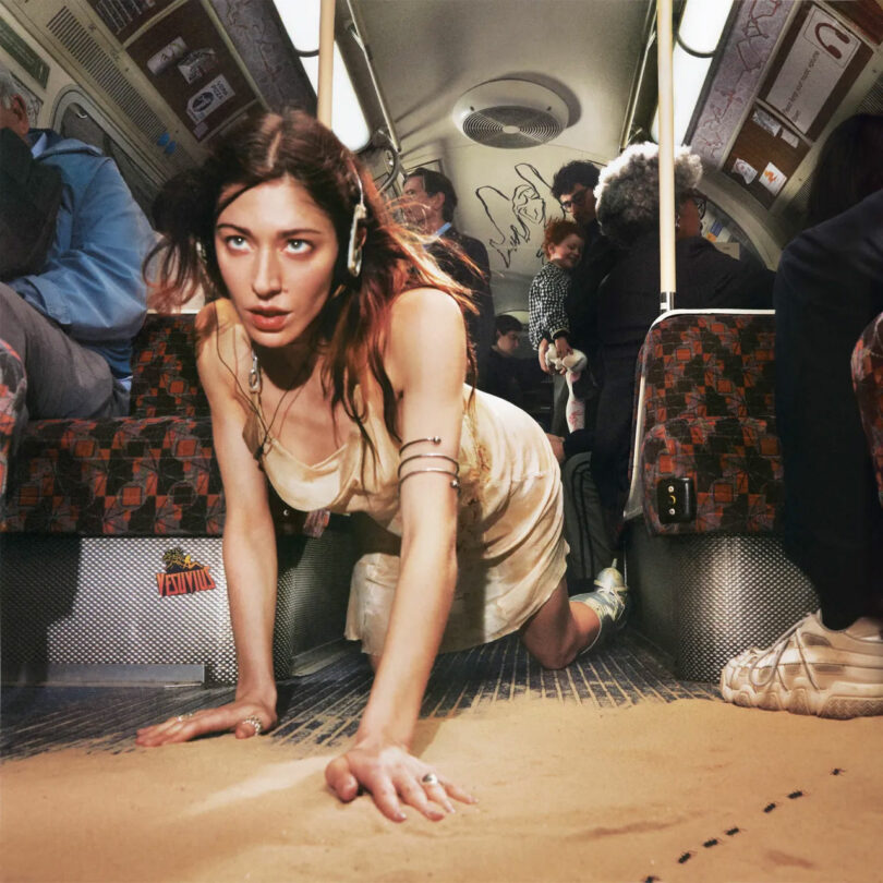 A woman in a dress and headphones crawls on a sand-covered aisle inside a subway train. Passengers are seated on both sides. The atmosphere is surreal.