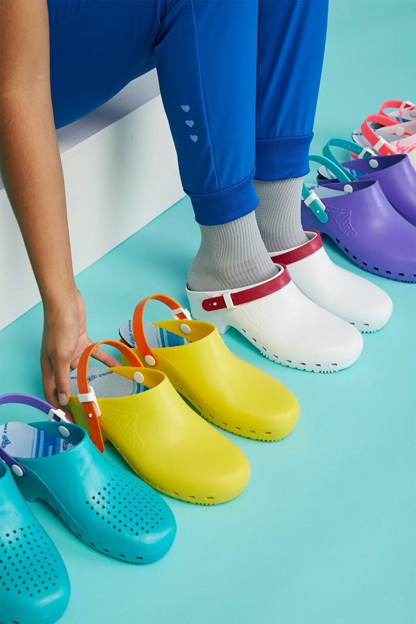 Person seated, adjusting a pair of teal clogs among a row of colorful clogs on a turquoise floor.