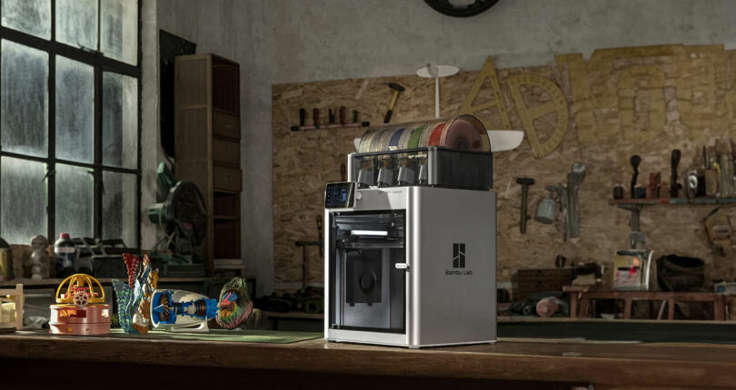 A 3D printer sits on a wooden table in a workshop, surrounded by colorful printed objects. A large window and various tools are visible in the background.