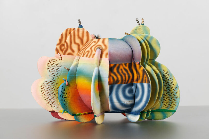 A colorful abstract sculpture made of layered, patterned shapes in various hues, including blue, orange, and pink, displayed against a plain background.