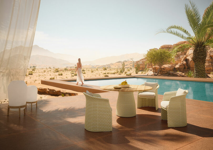 On the serene outdoor desert patio, a sleek DEDON table cradles a bowl of fruit. Nearby, four chairs invite relaxation beside the shimmering pool. A person stands contemplatively by the water with majestic mountains and clear skies painting the perfect backdrop.