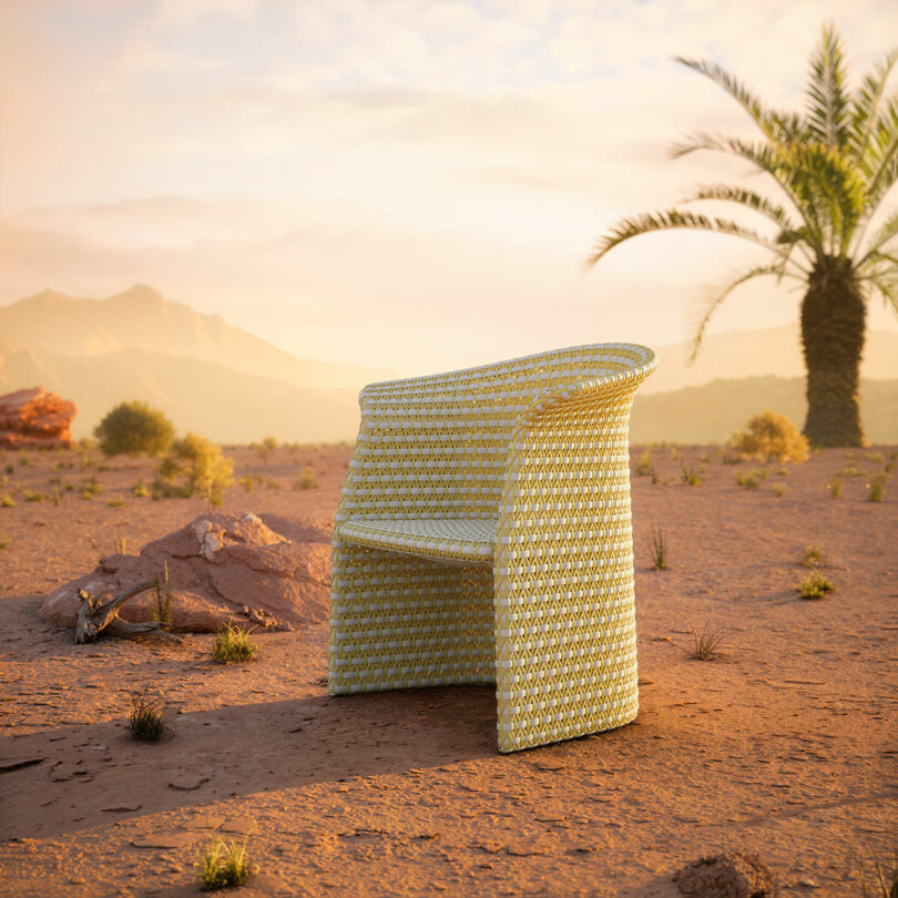 A DEDON wicker chair stands on a sandy desert landscape with scattered rocks and a palm tree, under a clear sky framing the distant mountains.