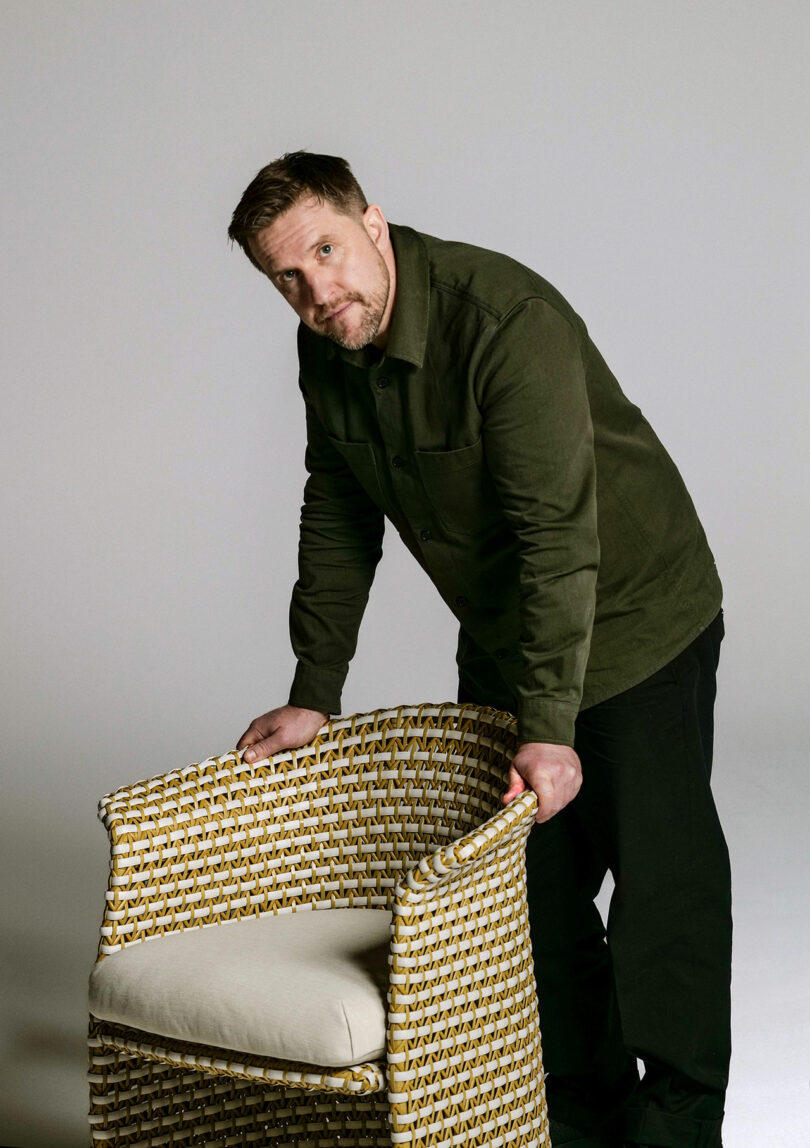 A person in a green jacket and black pants leans on the back of a cushioned wicker chair, reminiscent of DEDON's elegant style, against a plain background.