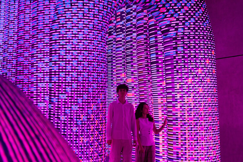 Two people stand inside a room with walls covered in a mosaic of pink, purple, and blue LED lights, creating a vibrant, immersive environment.