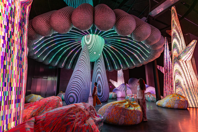 Une installation artistique colorée et immersive avec de grandes structures textiles illuminées ressemblant aux champignons et aux plantes. Deux personnes se tiennent parmi des sacs de haricots dynamiques, admirant l'affichage.