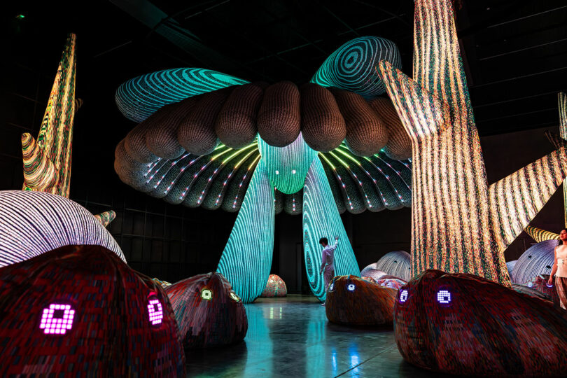 A person stands beneath a large, colorful, abstract mushroom-like light installation in a dimly lit room, surrounded by other illuminated structures.