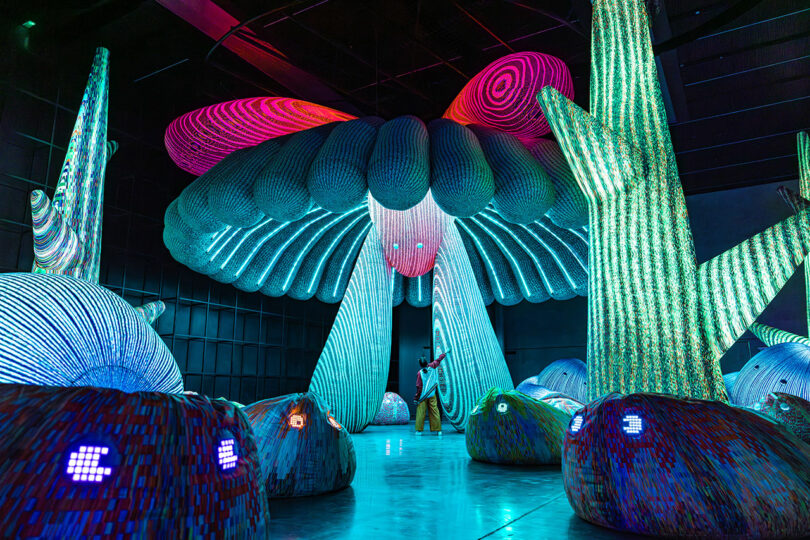 A person stands inside a vivid, large-scale art installation with colorful, illuminated, abstract structures resembling mushrooms and organic shapes.