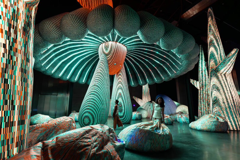 Une personne se tient dans une installation d'art immersive avec de grandes structures de champignons et de cactus brillantes avec des motifs rayés, entourés de sièges de coussin.