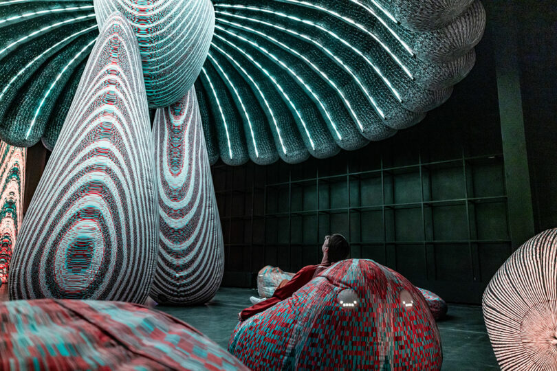 Une personne se trouve sous une grande installation abstraite éclairée avec des motifs tourbillonnants, entourés de grands coussins à motifs dans une pièce faiblement éclairée.