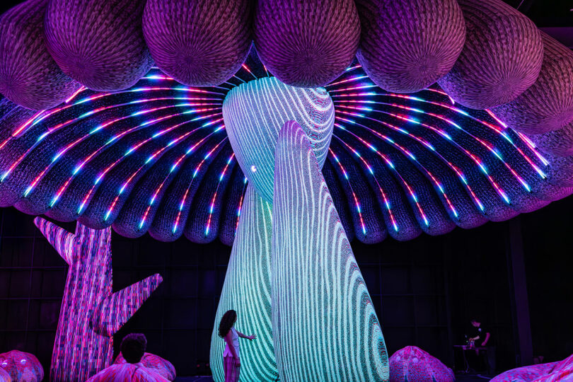 Une personne se tient sous une grande installation artistique interactive éclairée ressemblant à un champignon avec des lumières LED multicolores dans une pièce sombre.