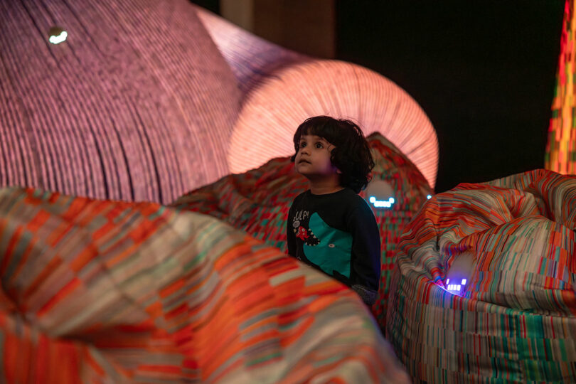 Child looking up, surrounded by colorful, illuminated fabric structures in a dark setting.