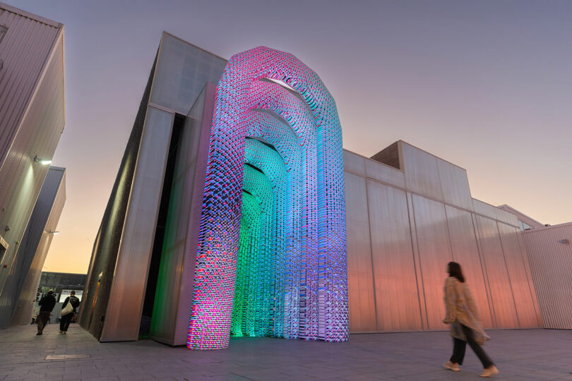 Une personne passe devant une grande installation artistique éclairée avec des couleurs vibrantes et des formes d'arc à l'extérieur des bâtiments modernes au crépuscule.