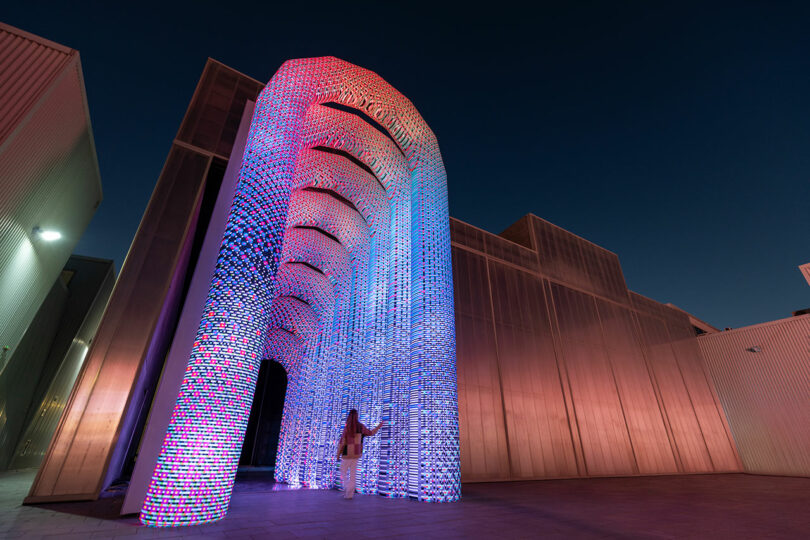 La personne se tient sous une grande arche illuminée avec des lumières rouges et bleues contre un ciel sombre.