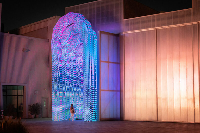 Une personne se dresse sous une grande structure en forme d'arc éclairée de lumières bleues et violettes, situées à l'extérieur près des bâtiments vêtus de métal.