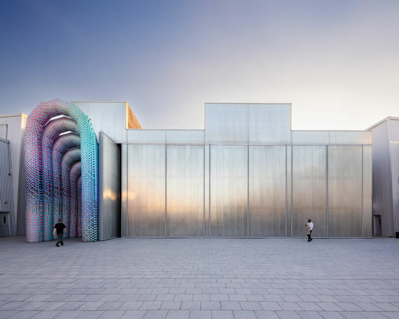 Modern building with a metallic facade, featuring a colorful arched structure on the left. Two people are walking nearby on a paved surface under a clear sky.