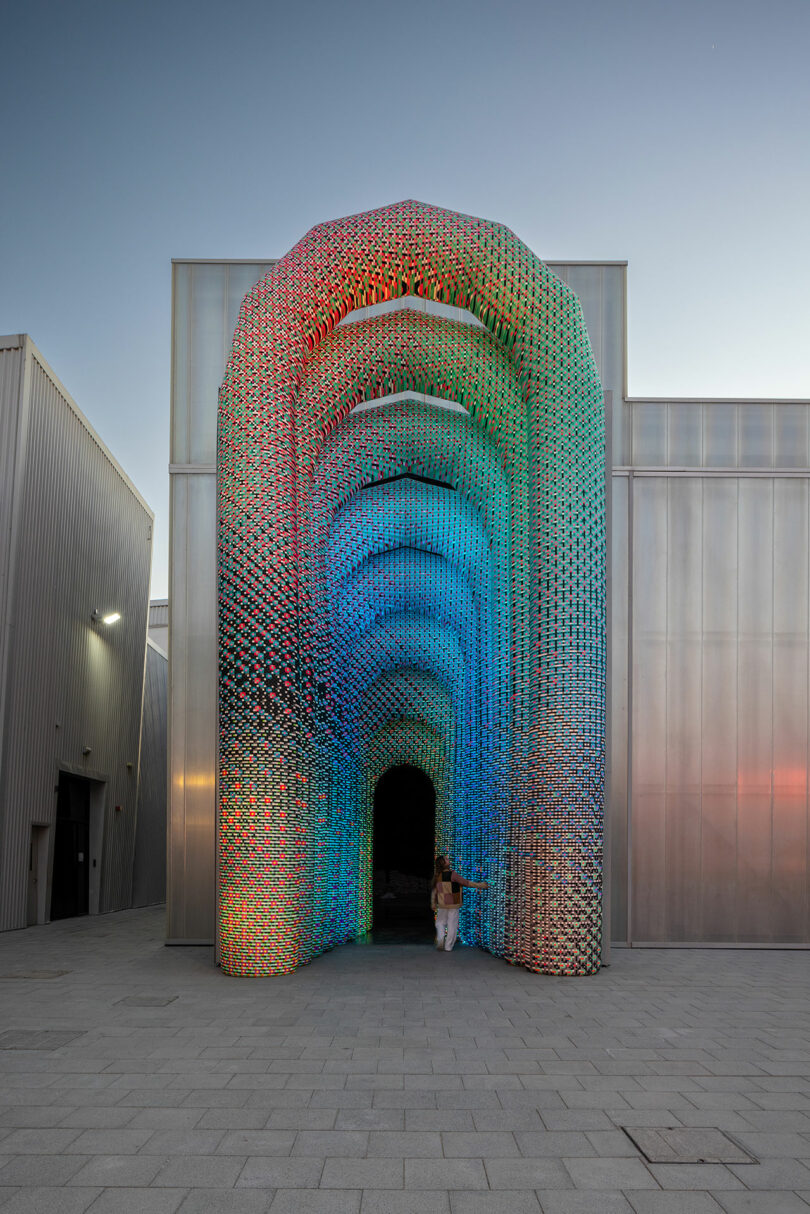 A person stands under a large, colorful, arch-shaped structure made of interlocking patterns. The building is gray with a metallic finish, and the sky is clear.