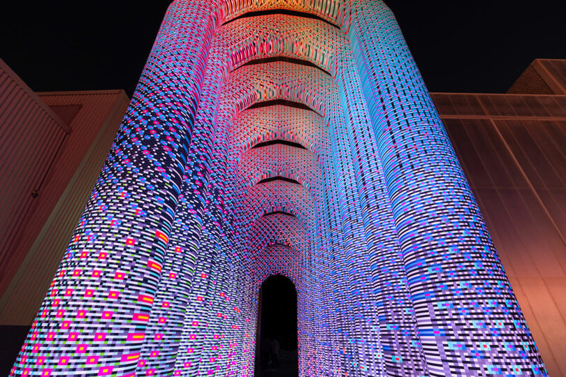 Un grand bâtiment cylindrique recouvert de motifs lumineux LED colorés et abstraits contre un ciel sombre.