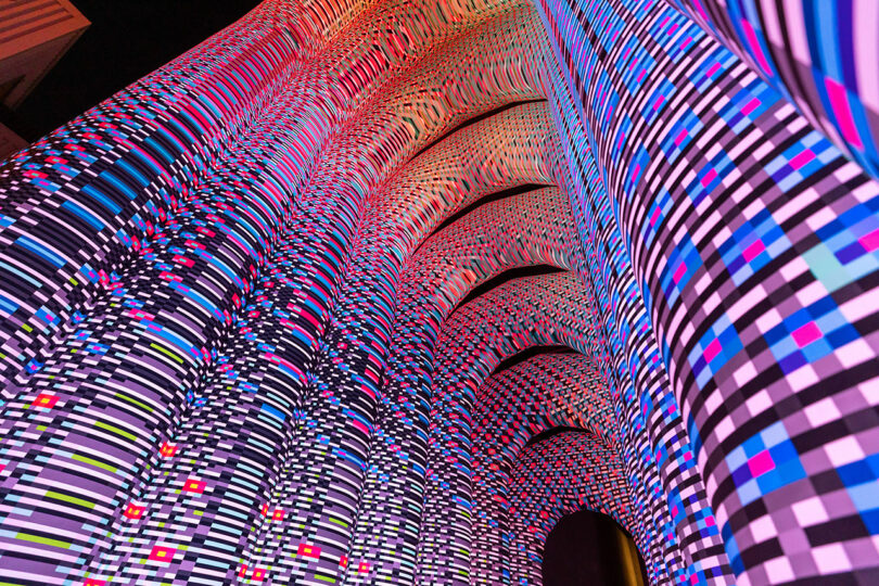 Vue verticale d'une façade de bâtiment illuminée de motifs lumineux colorés et pixelisés, créant un effet mosaïque numérique abstrait.