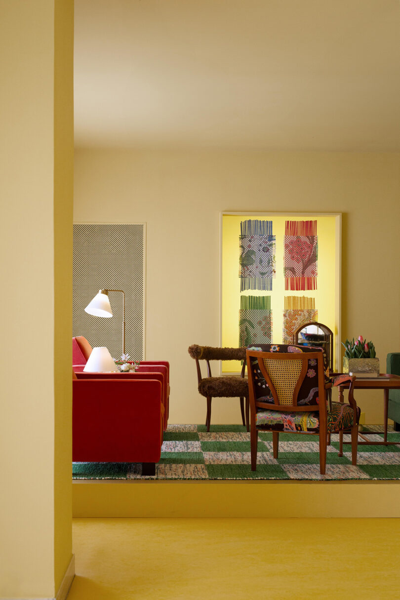 A room with red chairs, a wooden chair, and a coffee table. A standing lamp is on the left. Colorful artwork by Frank in the Loom is displayed in a backlit alcove. The floor is yellow and checkered green.