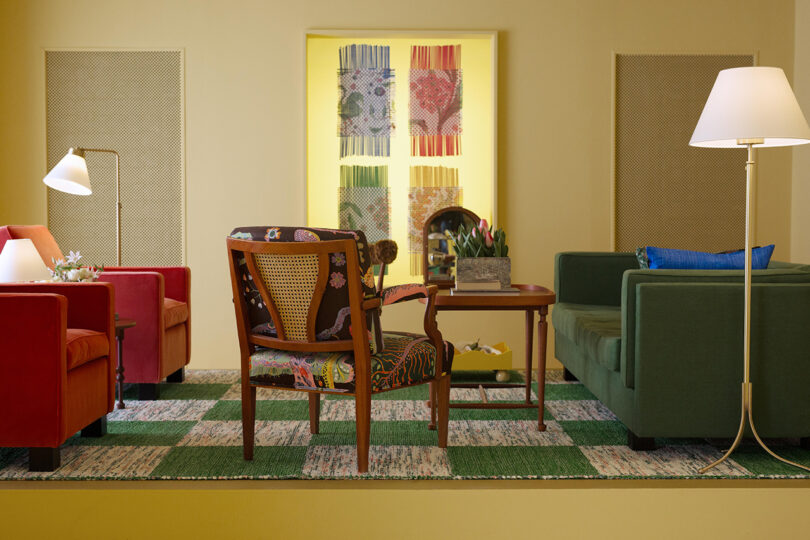 Seating arrangement with red and green chairs, a patterned wooden chair, and a wooden table. Two floor lamps illuminate the space. A colorful artwork hangs on the back wall.