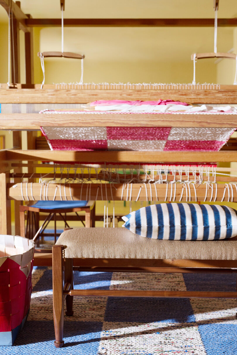 A wooden loom with partially woven fabric and a striped cushion in a brightly lit room. A basket is on the floor nearby.