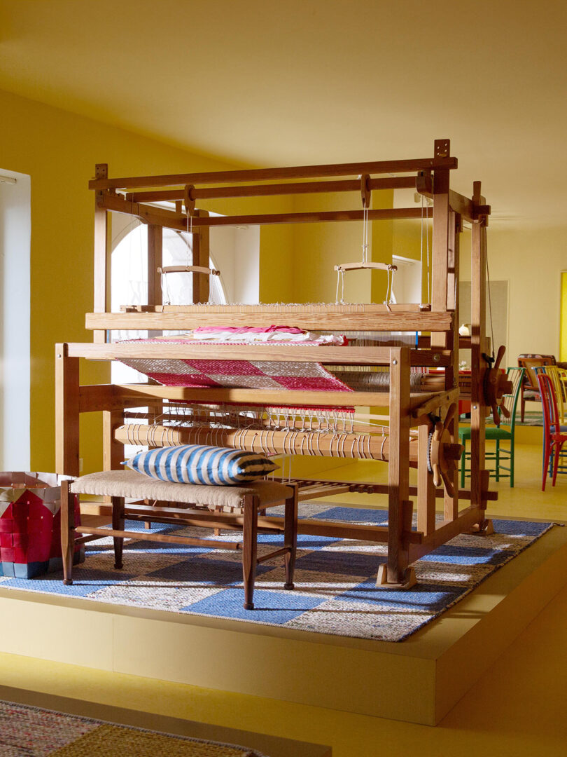 A wooden loom in a well-lit room with colorful fabric samples. The floor features a checkerboard rug pattern in shades of blue and yellow.